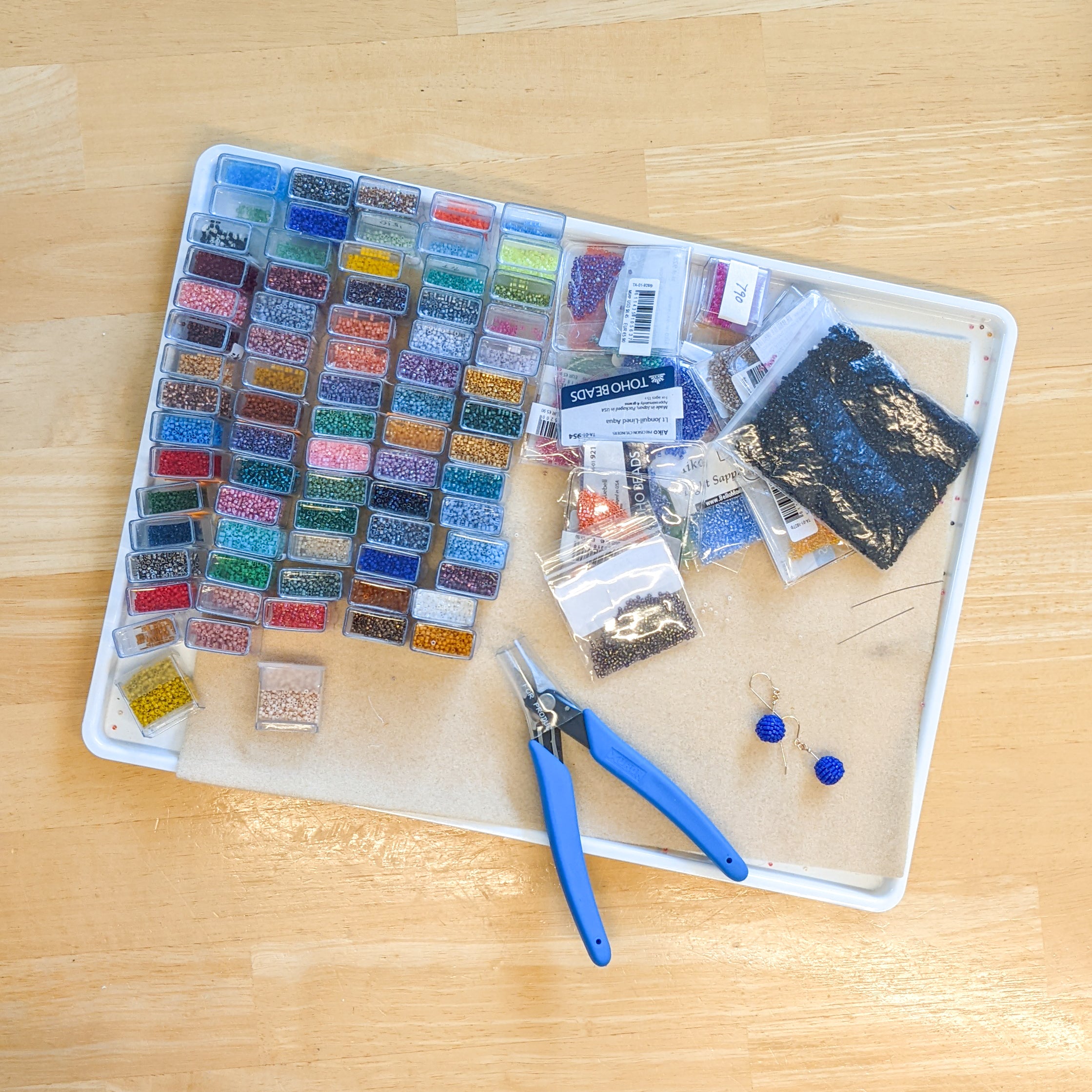 Many small rectangular flip top containers and several clear plastic pouches of seed beads, a pair of jewelry cutters, and a small pair of blue earrings on a matted tray